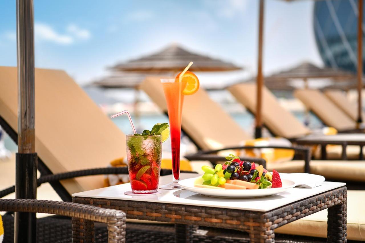 Al Raha Beach Resort & Spa Abu Dhabi Exterior foto The photo shows a table set outdoors, featuring two colorful cocktails and a plate of food. One cocktail is a vibrant red drink garnished with mint, while the other is a layered orange beverage with an orange slice on the rim. The plate of food inclu