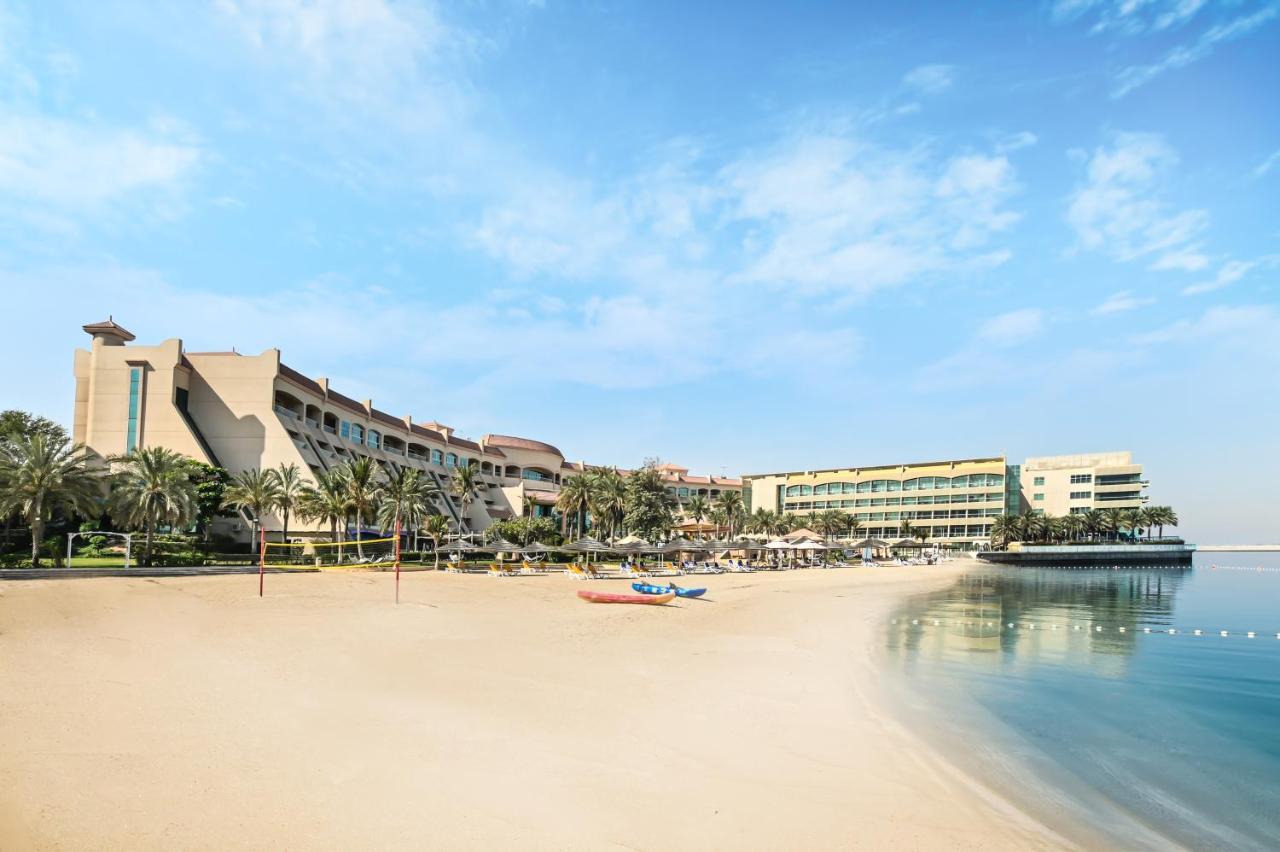 Al Raha Beach Resort & Spa Abu Dhabi Exterior foto The photo depicts a serene beach scene with a clear blue sky. In the foreground, there is a sandy beach that gently slopes down to calm water. On the left side, several palm trees provide a tropical atmosphere. In the background, there are two large 