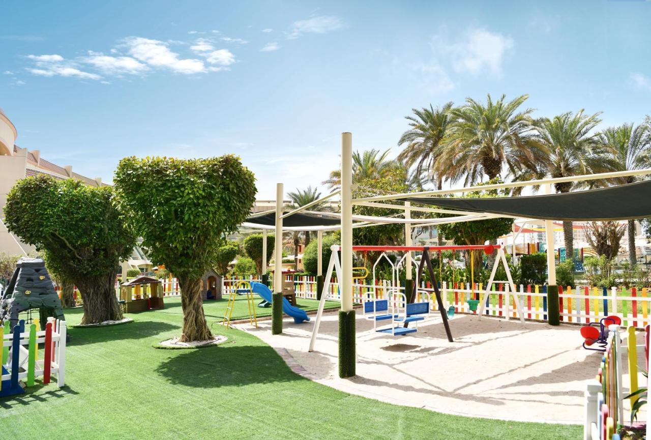 Al Raha Beach Resort & Spa Abu Dhabi Exterior foto The photo shows a brightly lit outdoor playground surrounded by palm trees. The playground features a variety of play equipment, including swings, a slide, and a climbing structure. The ground is covered in lush green artificial grass, providing a so