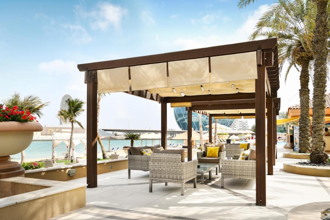 Al Raha Beach Resort & Spa Abu Dhabi Exterior foto The photo shows a stylish outdoor seating area by a beach. There is a wooden pergola covered with a light fabric shade, providing shelter to a set of comfortable wicker chairs and tables. The area is adorned with palm trees, and in the background, th
