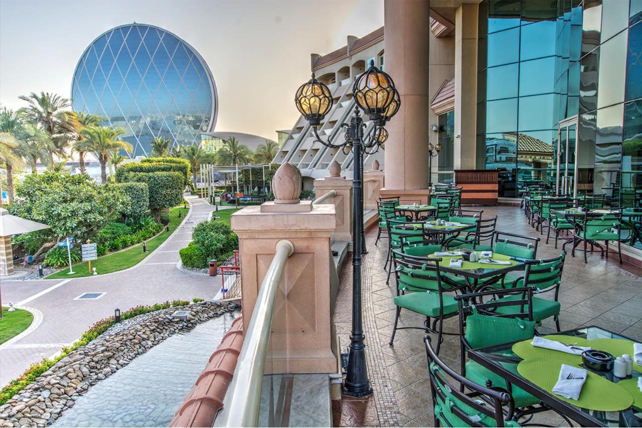 Al Raha Beach Resort & Spa Abu Dhabi Exterior foto The photo depicts an outdoor dining area with green tables and chairs arranged on a patio. In the background, there is a large, distinctive building with a striking blue dome, surrounded by well-maintained landscaping, including trees and plants. The