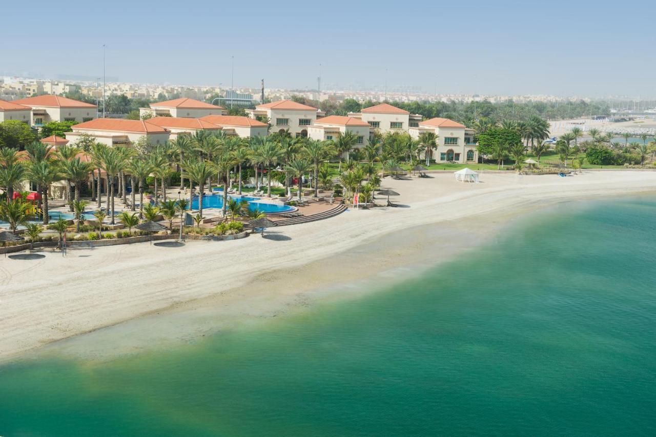 Al Raha Beach Resort & Spa Abu Dhabi Exterior foto The photo depicts a serene beach scene with a clear turquoise sea and a sandy shoreline. In the background, there are several buildings featuring a Mediterranean architectural style with red-tiled roofs. The area is adorned with palm trees and outdoo