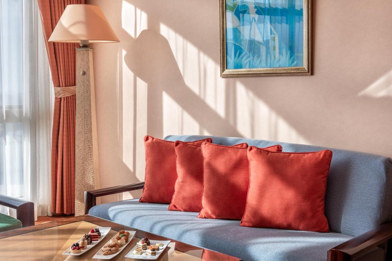 Al Raha Beach Resort & Spa Abu Dhabi Exterior foto The photo shows a cozy interior setting, likely a sitting area. It features a light blue couch with four red cushions. In front of the couch, there is a glass table displaying an assortment of snacks or small dishes. The room has soft lighting, with 