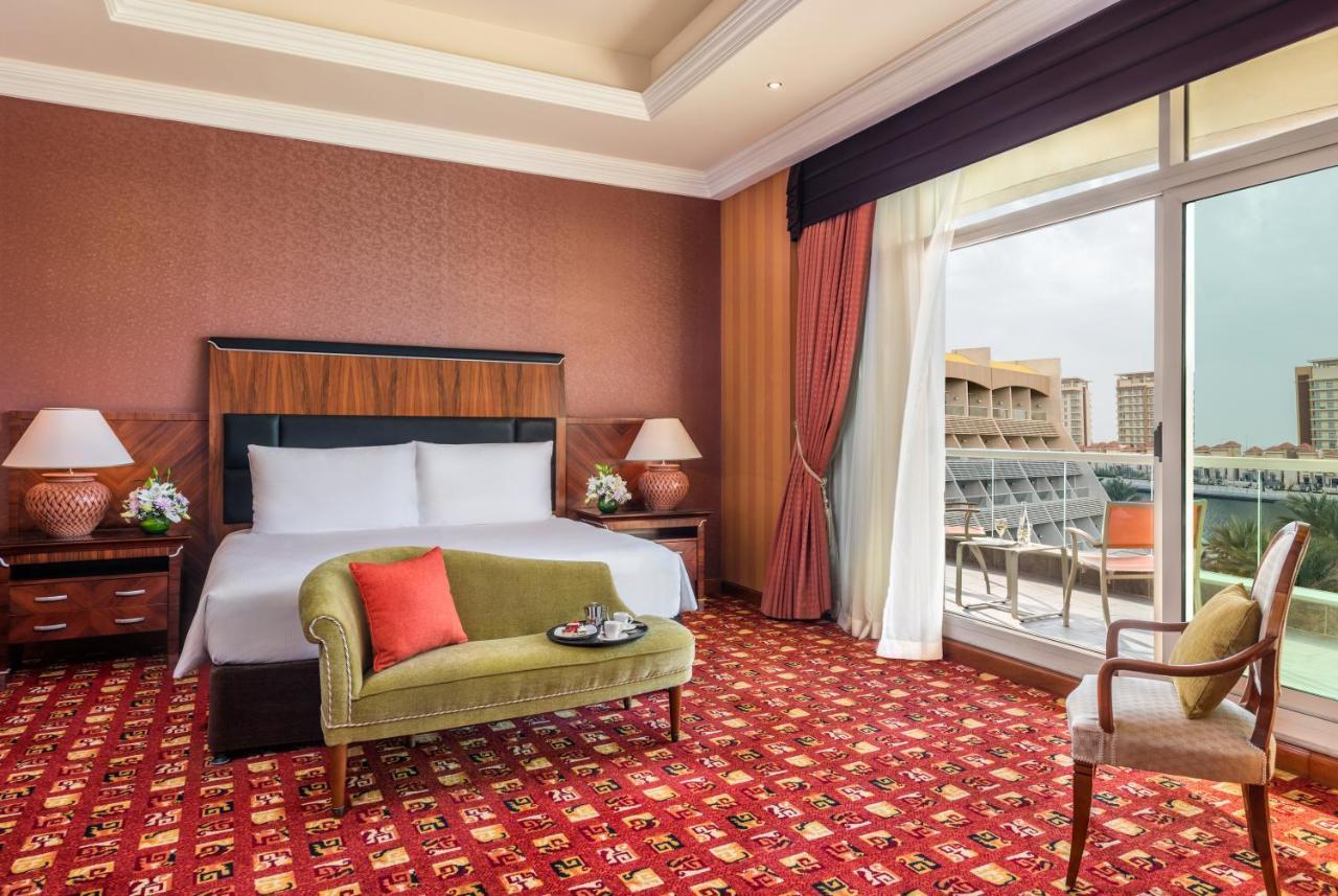 Al Raha Beach Resort & Spa Abu Dhabi Exterior foto The photo shows a hotel room with an elegant and modern design. It features a large bed with white linens and a dark wooden headboard. On either side of the bed are matching bedside lamps. There is a green armchair with a red pillow positioned near t