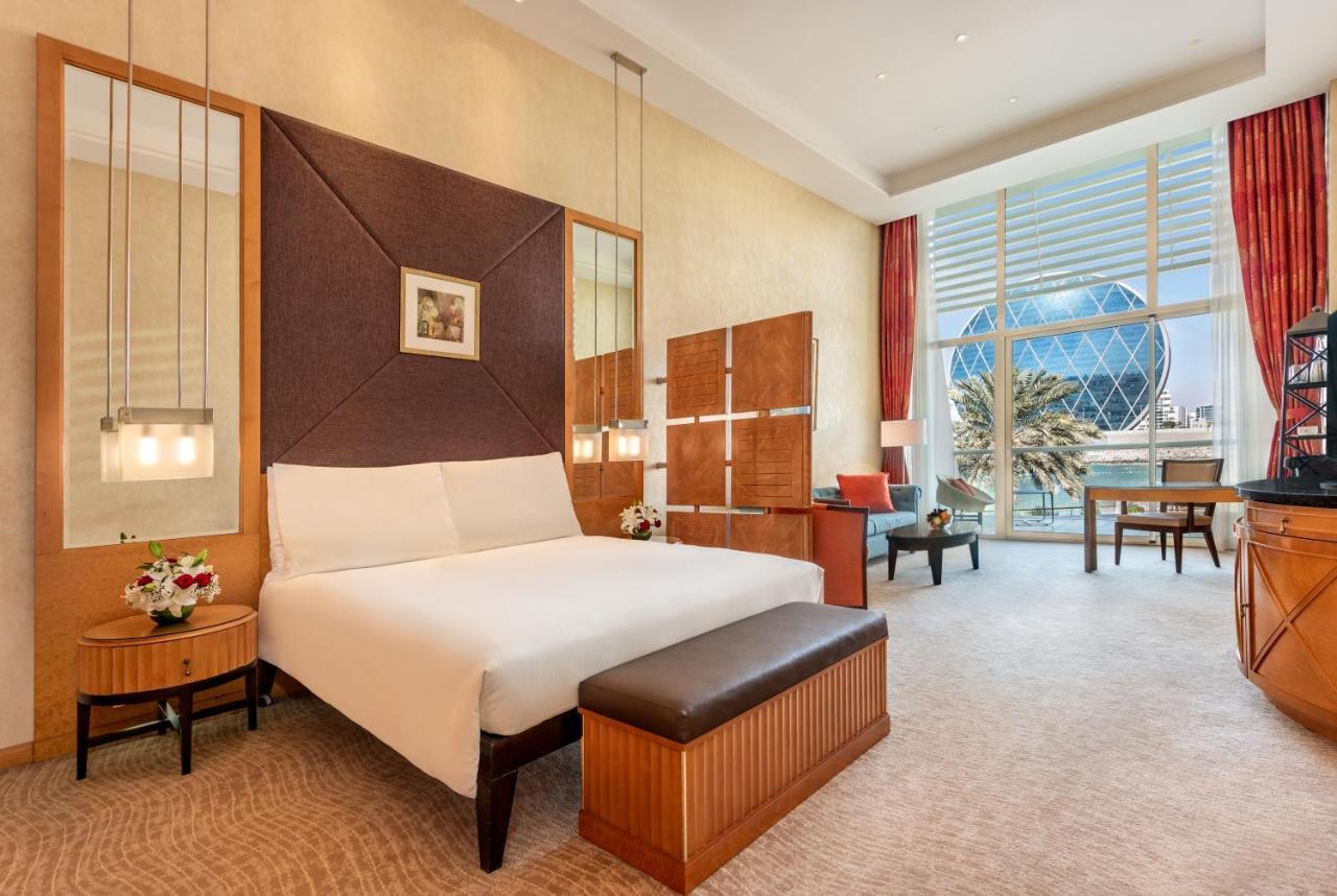 Al Raha Beach Resort & Spa Abu Dhabi Exterior foto The photo shows a modern hotel room featuring a neatly made bed with a white comforter, situated against a stylish headboard with a geometric design. There are bedside tables on either side of the bed, each with a small flower arrangement. A comforta