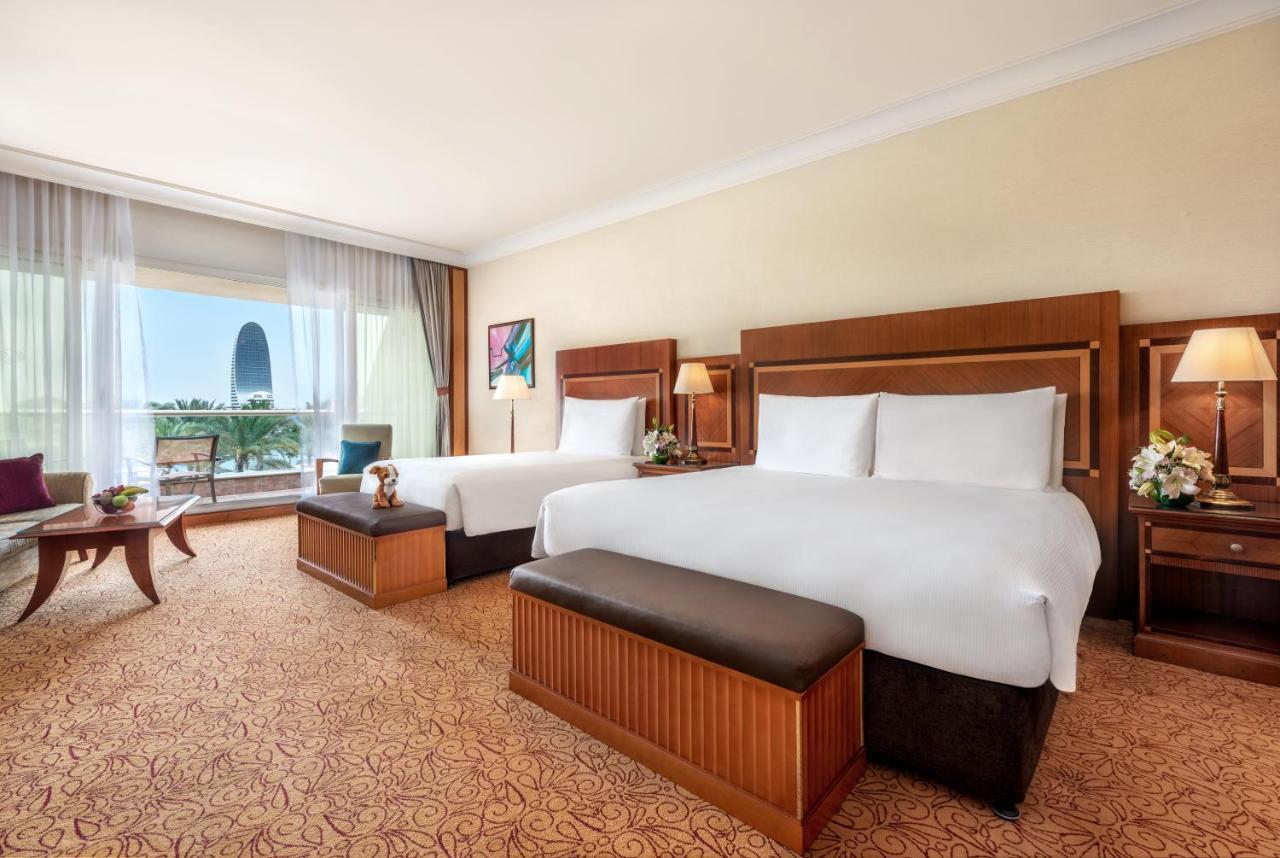 Al Raha Beach Resort & Spa Abu Dhabi Exterior foto The photo shows a spacious hotel room featuring two large beds with white linens and dark brown upholstered headboards. There are matching benches at the foot of each bed. The room is decorated with a warm color palette and has a carpeted floor. To o