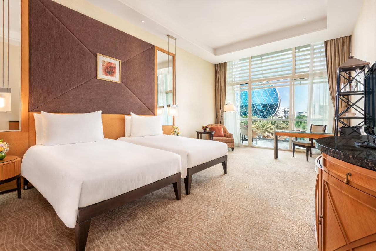 Al Raha Beach Resort & Spa Abu Dhabi Exterior foto The photo shows a well-decorated hotel room featuring two single beds with white linens. The room has a modern aesthetic, with a large window letting in natural light and offering a view of a circular structure outside, possibly a landmark or artisti