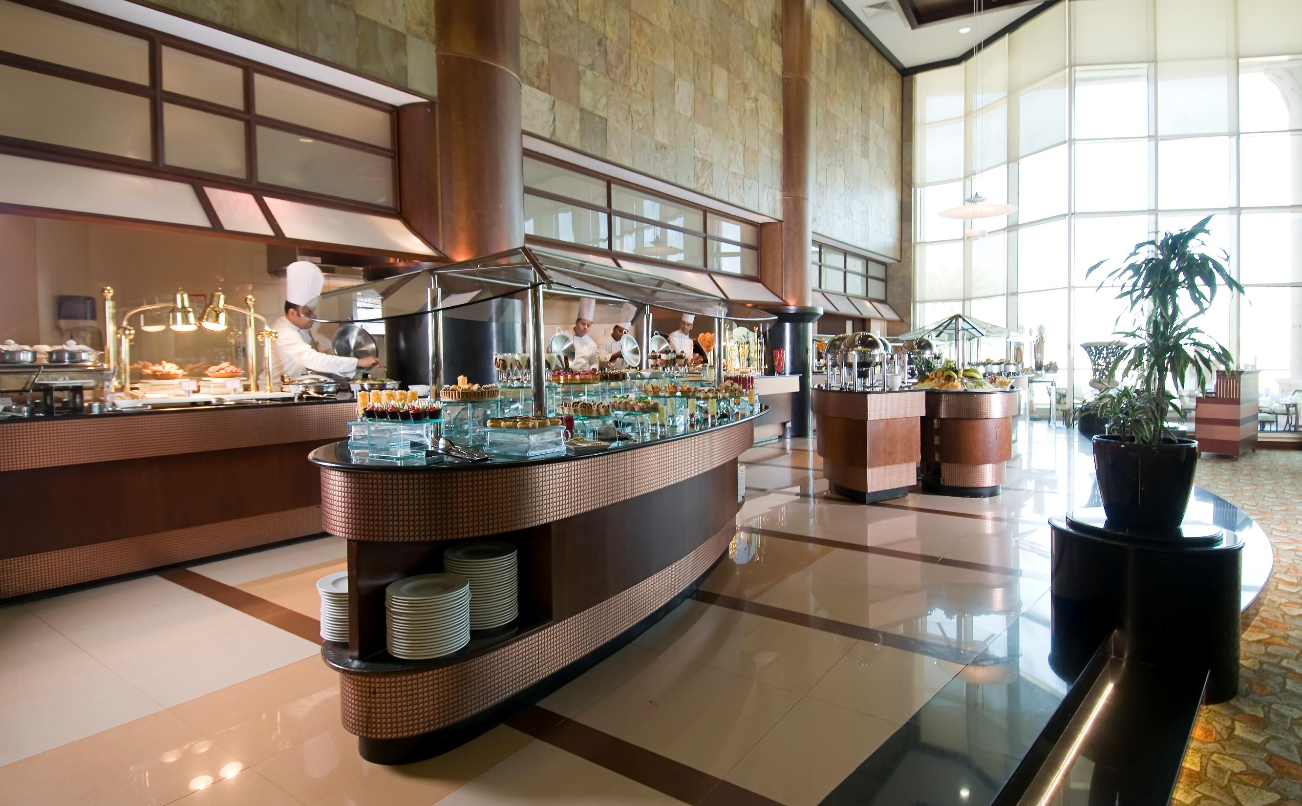 Al Raha Beach Resort & Spa Abu Dhabi Exterior foto The photo shows a spacious buffet area in a restaurant or hotel. There are multiple food stations with glass displays showcasing a variety of dishes. Chefs can be seen in the background, preparing food. The decor features large windows allowing natur