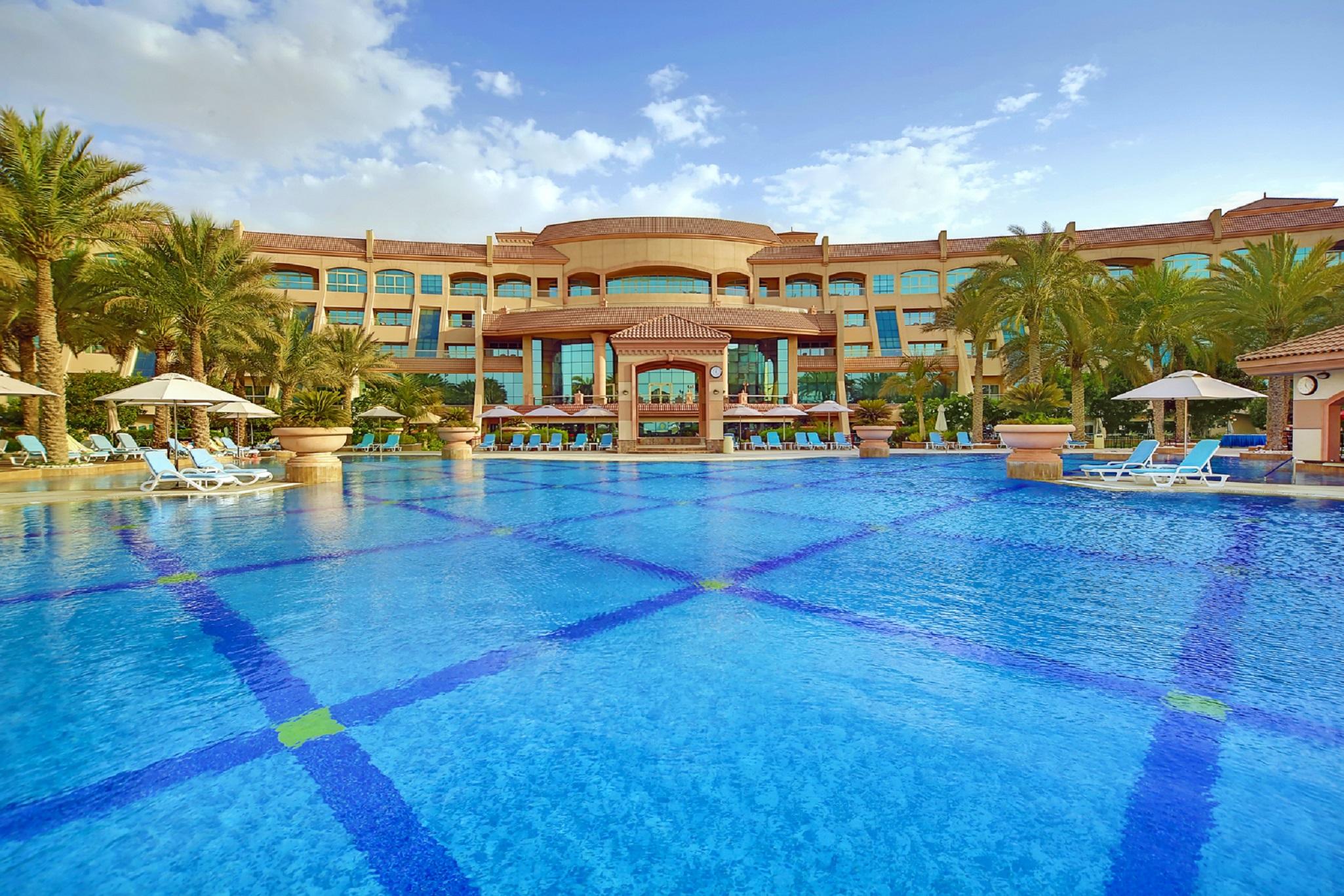 Al Raha Beach Resort & Spa Abu Dhabi Facilidades foto The photo features a luxurious resort with a large swimming pool in the foreground. The pool is crystal clear and reflects the sky, bordered by lounge chairs and palm trees. In the background, the resort building showcases modern architecture with la