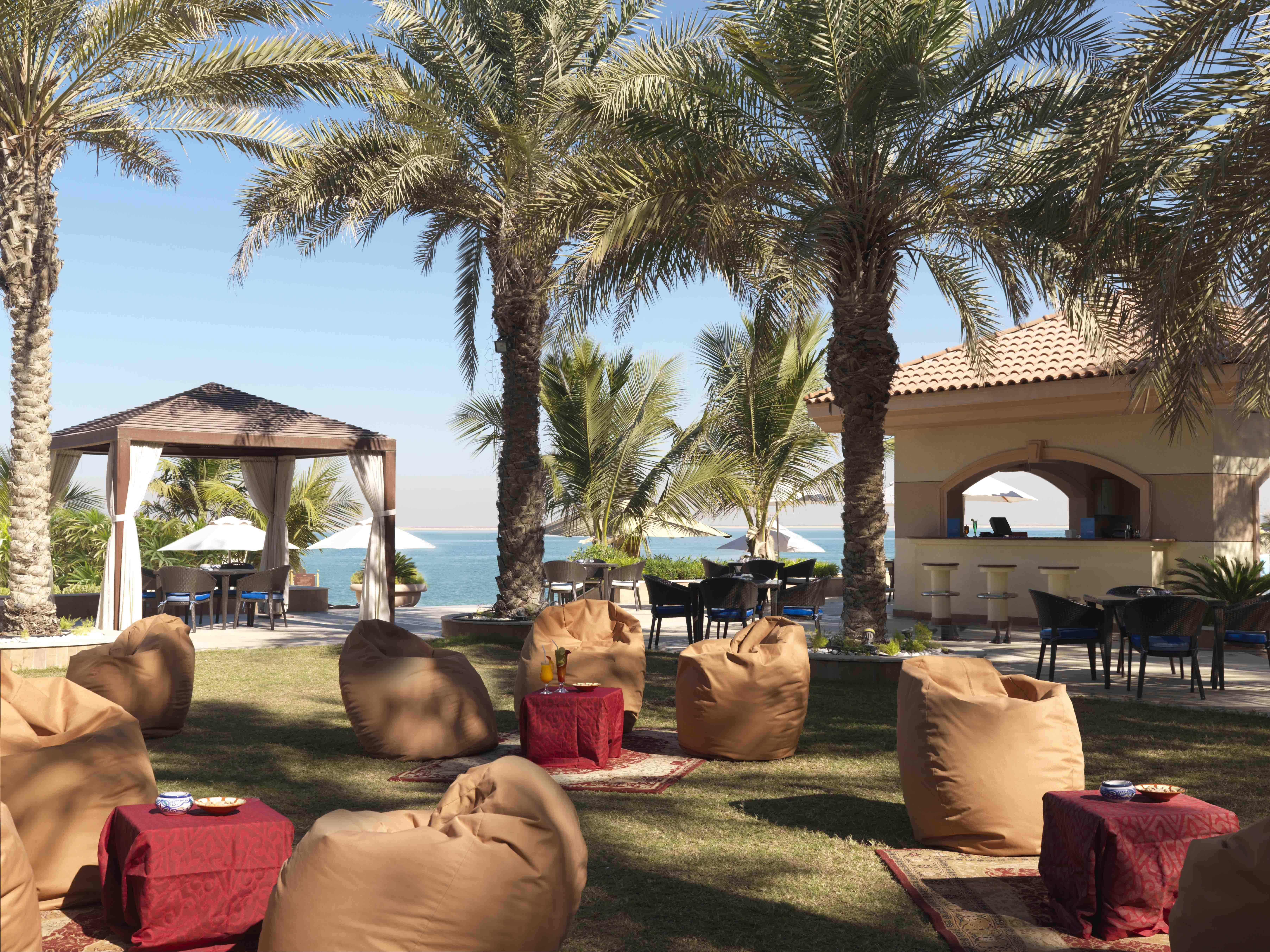 Al Raha Beach Resort & Spa Abu Dhabi Restaurante foto The photo depicts a beautiful outdoor setting with several elements of relaxation and leisure. In the foreground, there are large, comfortable bean bag chairs arranged on a grassy area, accompanied by low tables. Surrounding this seating area are tal