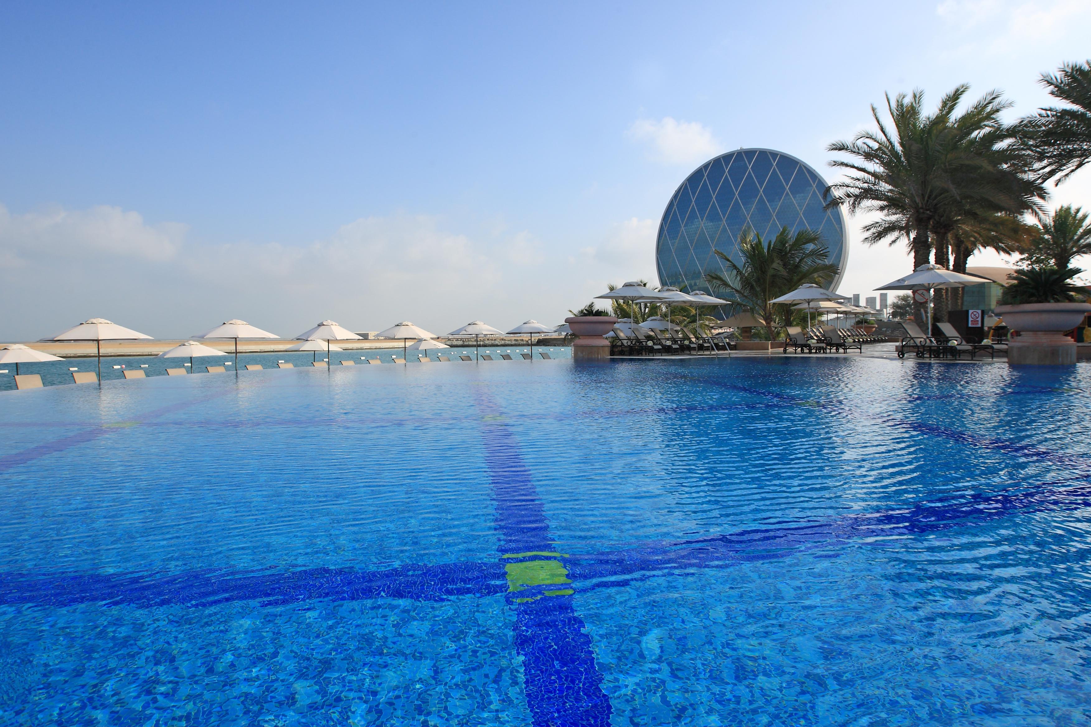 Al Raha Beach Resort & Spa Abu Dhabi Exterior foto The photo features a large, serene swimming pool with clear, blue water reflecting the sky. Surrounding the pool are several lounge chairs and umbrellas, suggesting a relaxed, resort-like atmosphere. In the background, a distinctive building with a r
