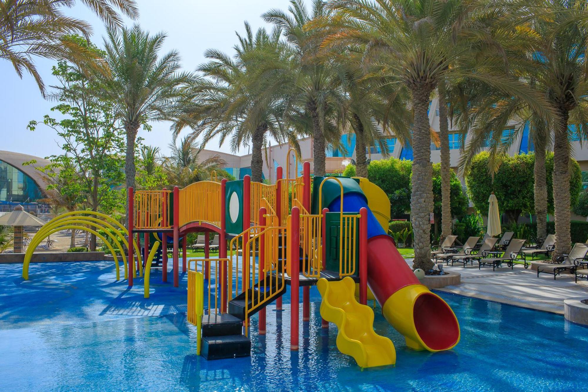 Al Raha Beach Resort & Spa Abu Dhabi Exterior foto The photo shows a colorful water play area located in a pool setting. It features a variety of play structures, including slides in multiple colors, such as yellow, red, and blue. Surrounding the play structure are palm trees, creating a tropical atm