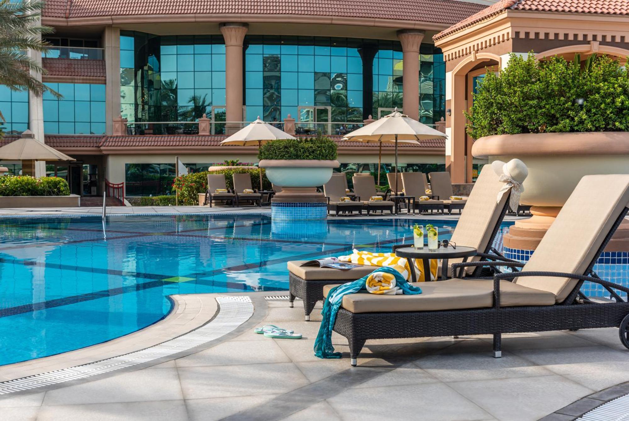 Al Raha Beach Resort & Spa Abu Dhabi Exterior foto The photo depicts a luxurious outdoor swimming pool area. In the foreground, there are two lounge chairs with cushions, positioned beside the pool. A small table between them holds drinks and snacks, suggesting a relaxing atmosphere. The pool itself 
