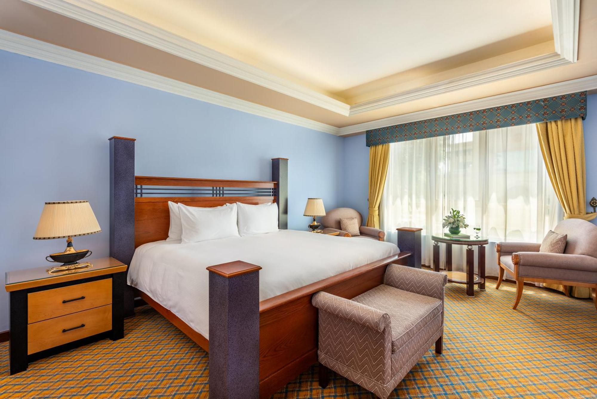 Al Raha Beach Resort & Spa Abu Dhabi Exterior foto The photo shows a well-decorated hotel room. It features a large bed with a wooden frame, neatly made with white bedding. On either side of the bed are bedside tables with lamps. There is a comfortable chair next to a window that has sheer curtains a