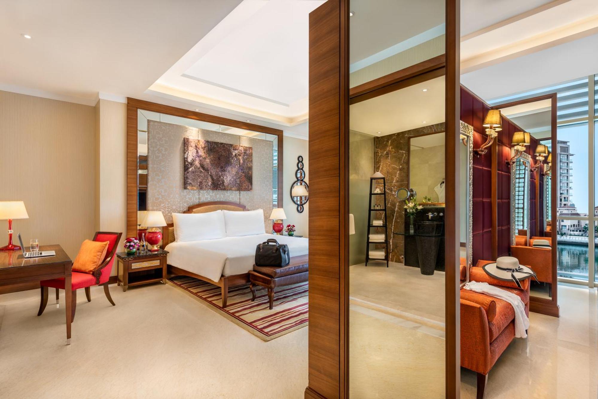 Al Raha Beach Resort & Spa Abu Dhabi Exterior foto The photo shows a luxurious hotel room with a modern design. The room features a large bed with white linens, flanked by bedside tables with lamps. There is a area with a chair and a small table on the left side, and a vibrant artwork hanging above t
