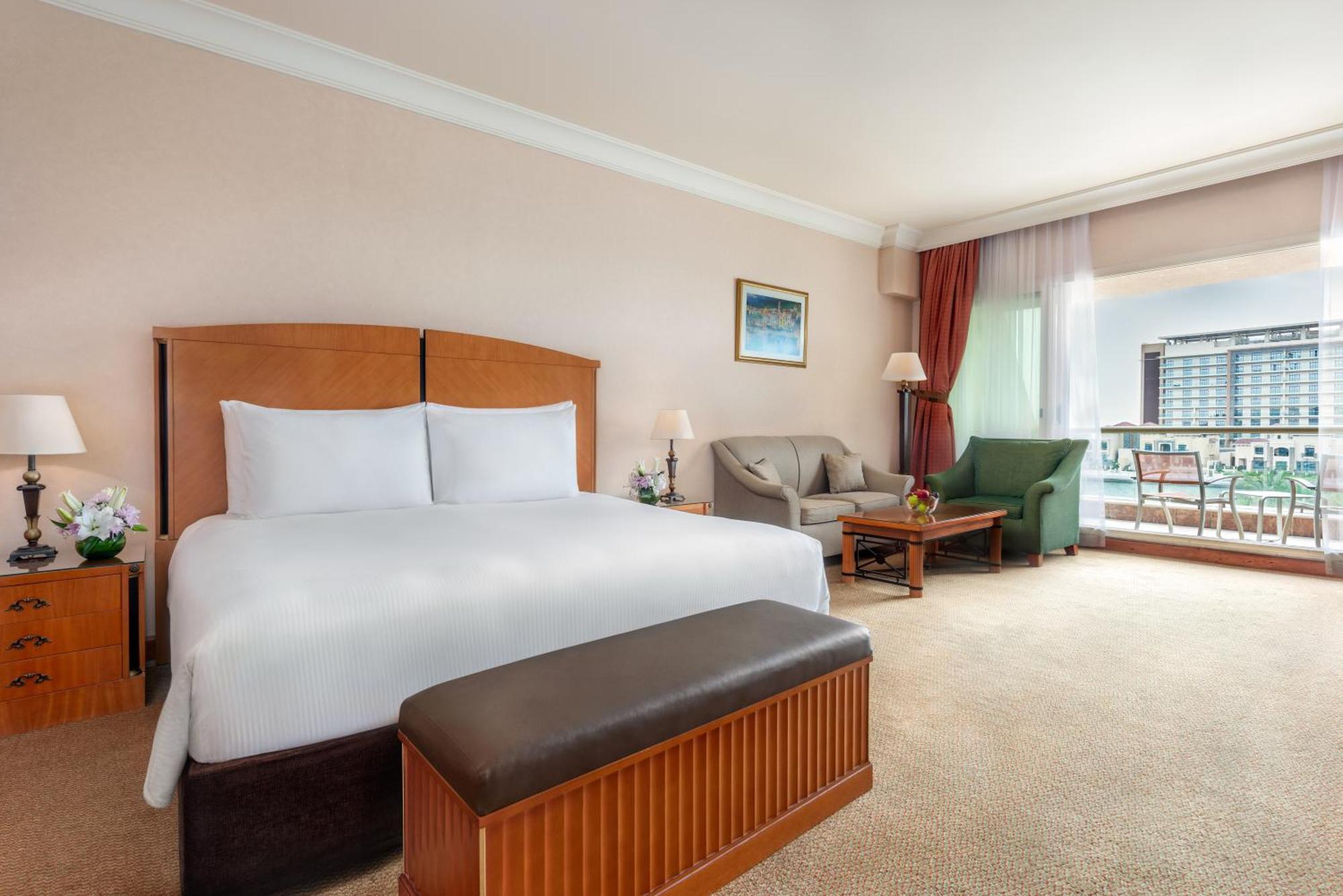 Al Raha Beach Resort & Spa Abu Dhabi Exterior foto The photo shows a well-furnished hotel room featuring a large bed with white linens and a wooden headboard. There's a brown bench at the foot of the bed. To one side, a sitting area includes a sofa and a coffee table, complemented by two lamps. Large