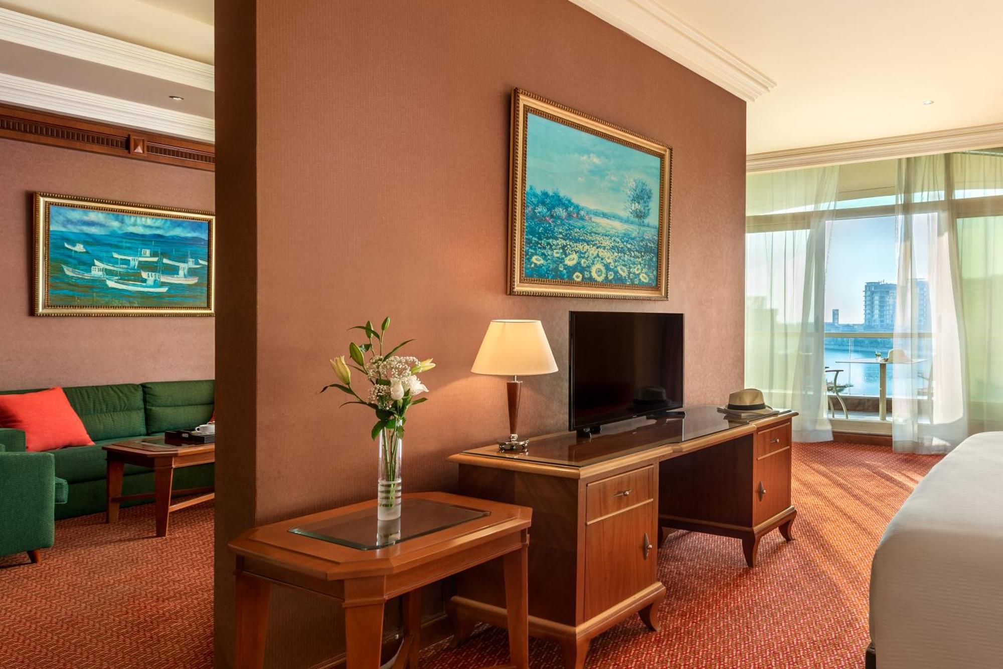 Al Raha Beach Resort & Spa Abu Dhabi Quarto foto The photo shows a hotel room with a warm, inviting atmosphere. There's a large window that provides natural light and a view, likely overlooking a cityscape or water. On one wall, there's a piece of artwork featuring a landscape, and nearby, a small 