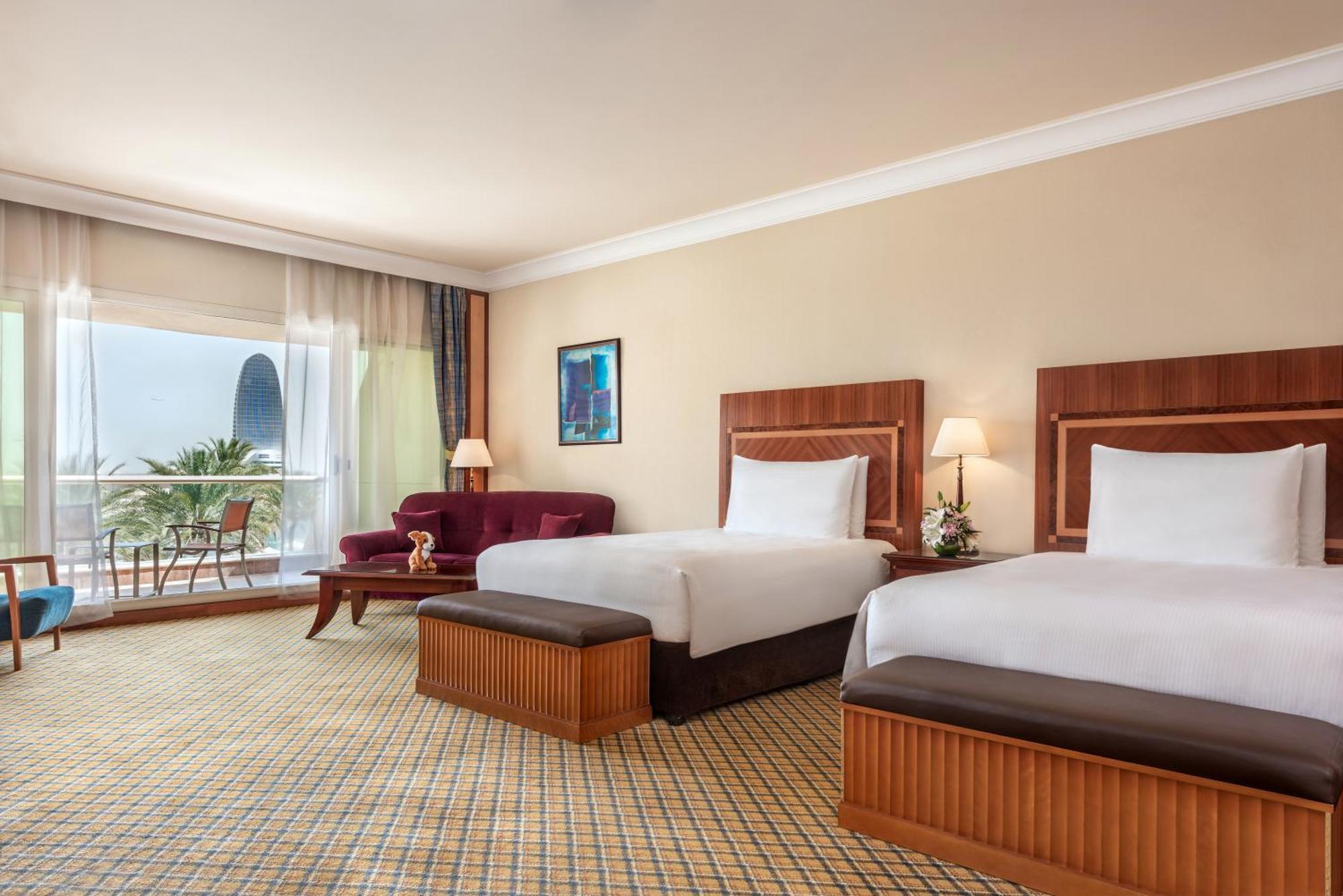 Al Raha Beach Resort & Spa Abu Dhabi Quarto foto The photo shows a hotel room featuring two twin beds with wooden headboards. The room has a light, neutral color scheme, with a large window that provides natural light and a view of the outdoors. There is a small seating area with a purple sofa and 