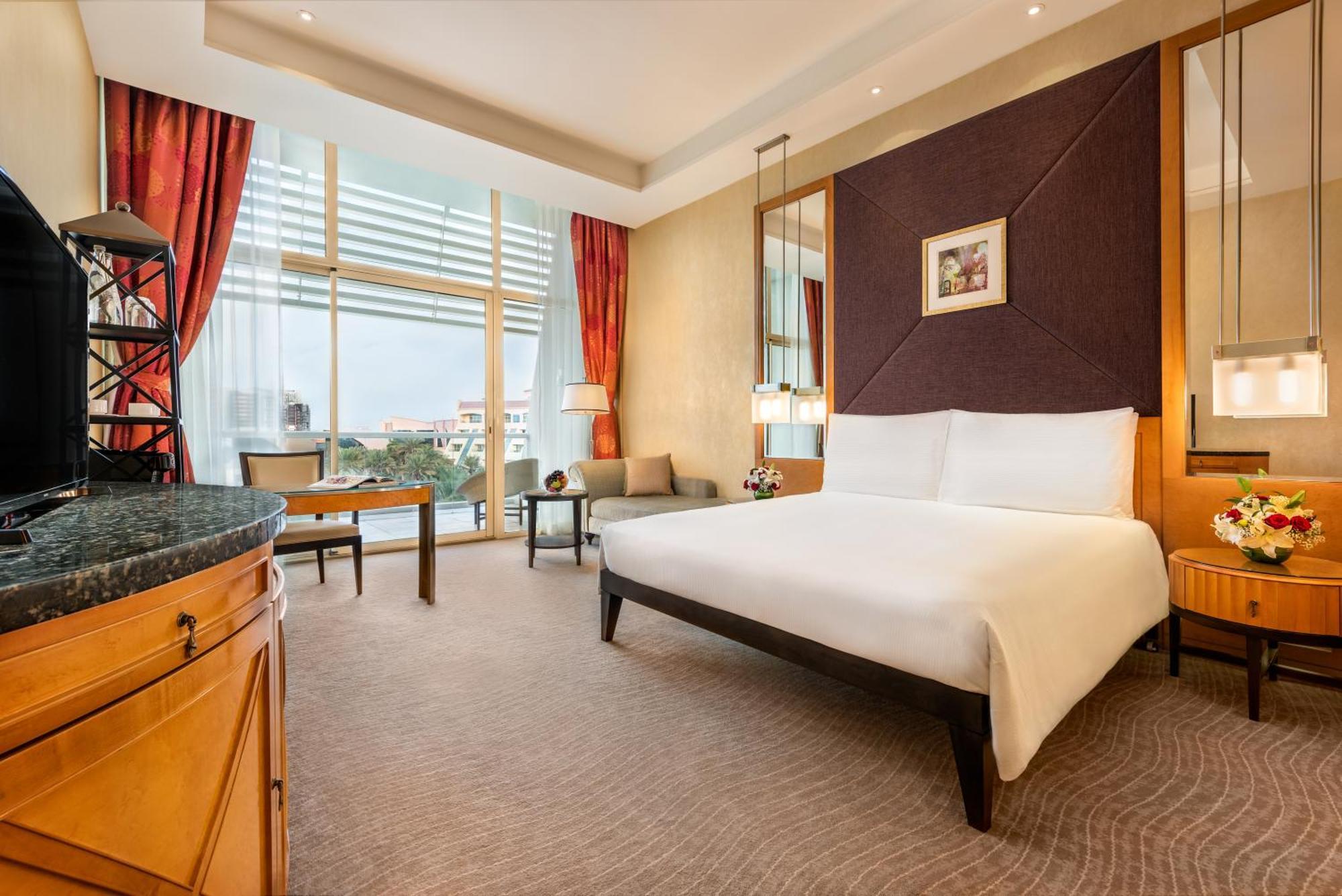Al Raha Beach Resort & Spa Abu Dhabi Quarto foto The photo shows a modern hotel room featuring a large bed with a white duvet positioned against a purple accent wall. There are tall windows with sheer curtains allowing natural light to enter the space, along with red drapes framing the windows. A d