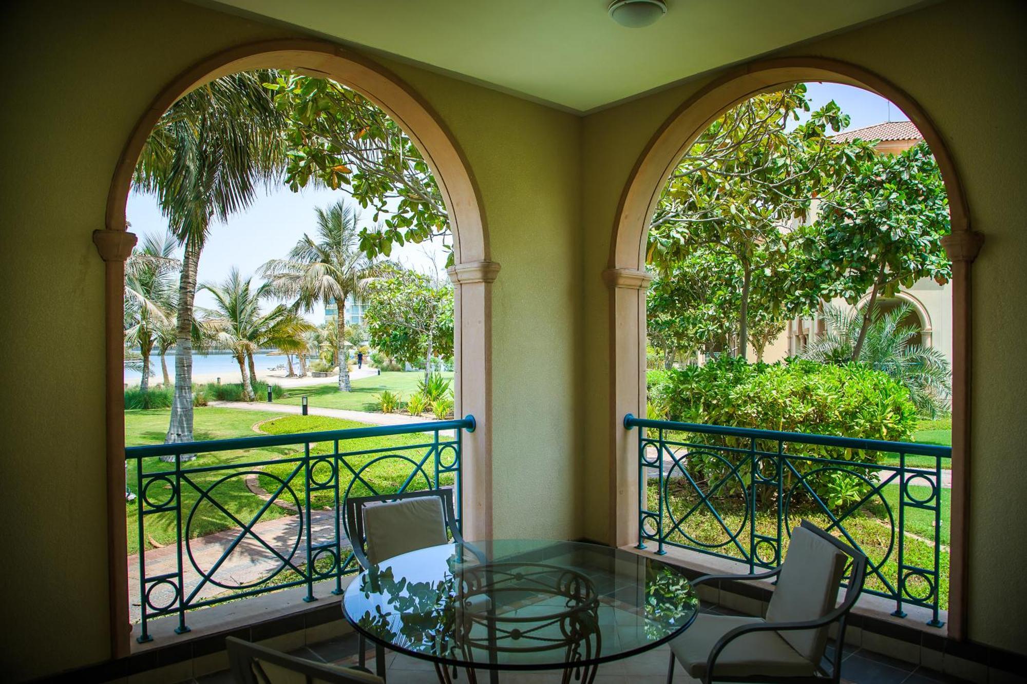 Al Raha Beach Resort & Spa Abu Dhabi Quarto foto The photo displays a beautifully designed balcony or terrace area. It features two large arches that frame the view outside. Through the arches, you can see a lush garden with palm trees and well-maintained greenery. There’s a glass table with severa