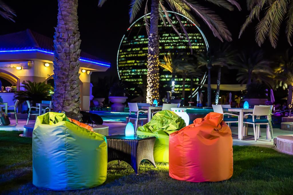 Al Raha Beach Resort & Spa Abu Dhabi Exterior foto The photo shows a vibrant outdoor lounge area at night. In the foreground, there are colorful bean bag chairs—one in bright green, one in yellow, and one in orange. A small table sits between the bean bags. In the background, illuminated palm trees a
