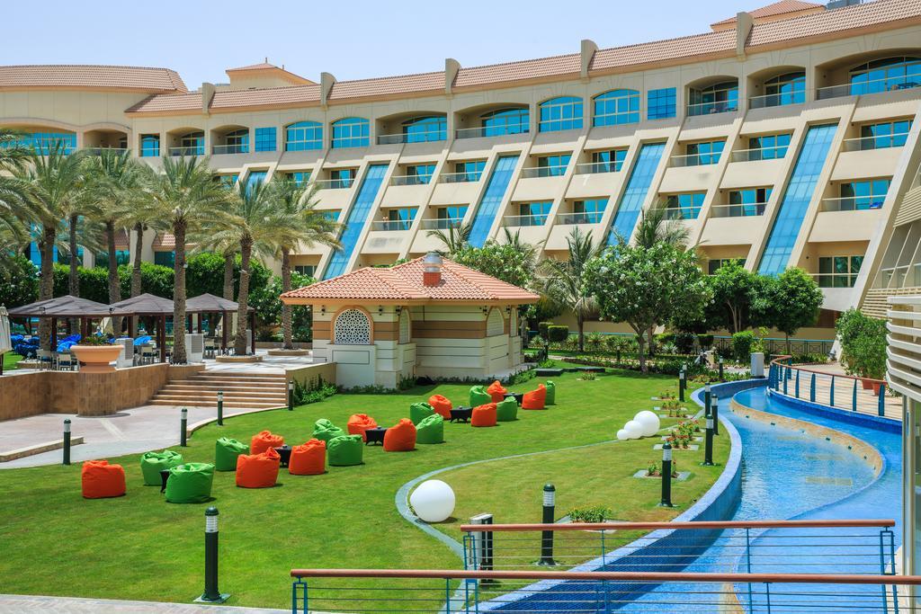 Al Raha Beach Resort & Spa Abu Dhabi Exterior foto The photo depicts a modern hotel complex with multiple stories and large windows. In the foreground, there is a well-maintained lawn featuring colorful lounge chairs in shades of orange and green. A pathway runs alongside a blue water feature, creati