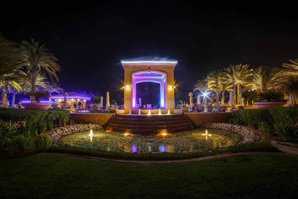 Al Raha Beach Resort & Spa Abu Dhabi Exterior foto The photo depicts a beautifully lit outdoor scene at night. In the foreground, there is a serene water feature with a circular pond, and small fountains creating gentle ripples. Surrounding the pond are lush green lawns and ornamental plants. 

In th