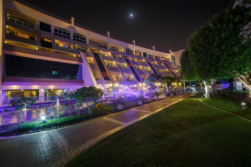Al Raha Beach Resort & Spa Abu Dhabi Exterior foto The photo shows a modern building illuminated at night, with a vibrant purple hue. The structure has multiple floors, each with balconies overlooking a landscaped area. In the foreground, there is a pathway lined with well-manicured grass and shrubs,