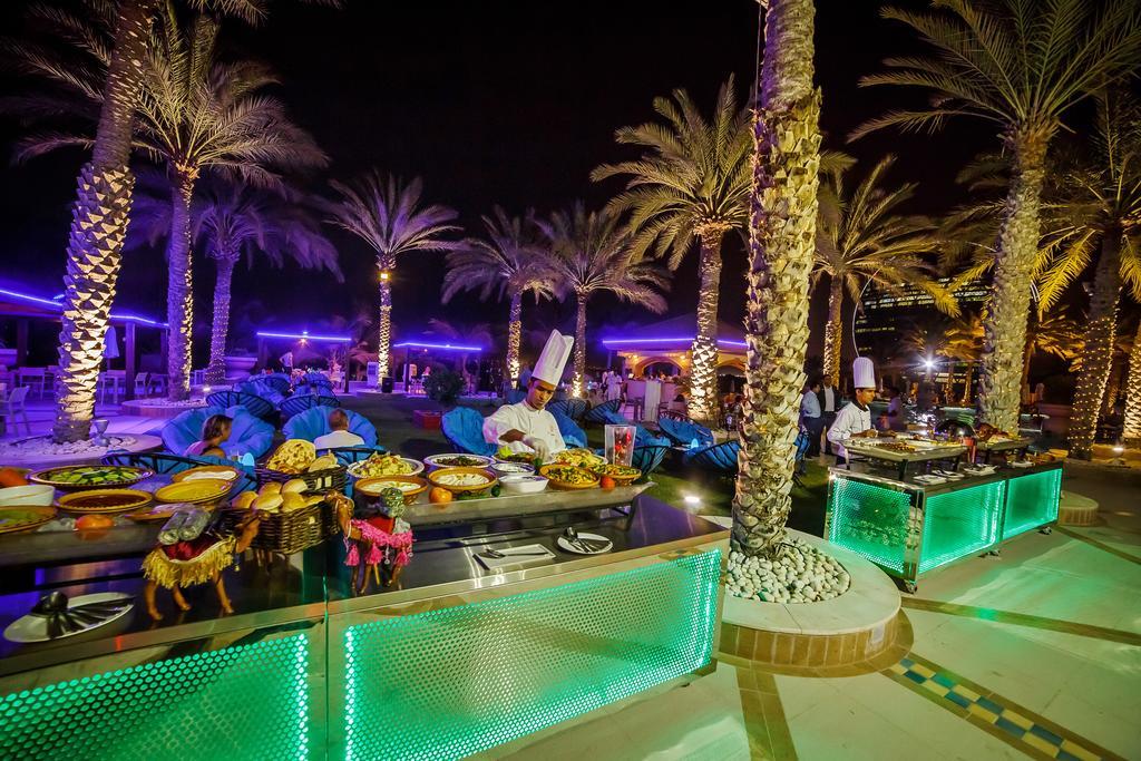 Al Raha Beach Resort & Spa Abu Dhabi Exterior foto The photo depicts an outdoor dining area at night, featuring several chefs in white uniforms and hats working at a food station. The setting is illuminated by colorful lights, with glowing green accents on the serving tables. Surrounding the area are